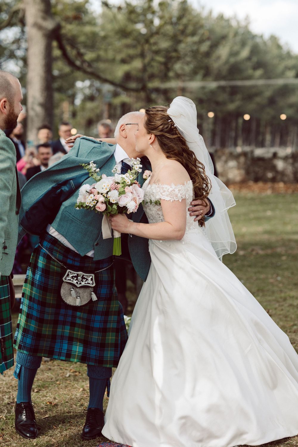 A bride and groom share a romantic moment outdoors while wearing traditional wedding attire with Scottish tartan details.