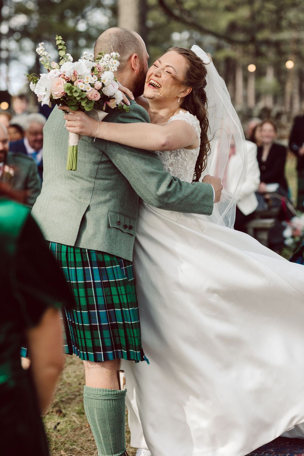 A joyful wedding moment captured outdoors with the groom in traditional Scottish kilt and bride in white dress sharing a laugh.