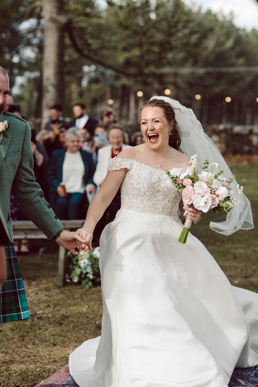 Bride in off-shoulder wedding gown laughs with pure joy during outdoor ceremony with Scottish-themed attire.