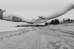 Black and white photo of couple reaching hands toward each other on sandy beach shoreline.
