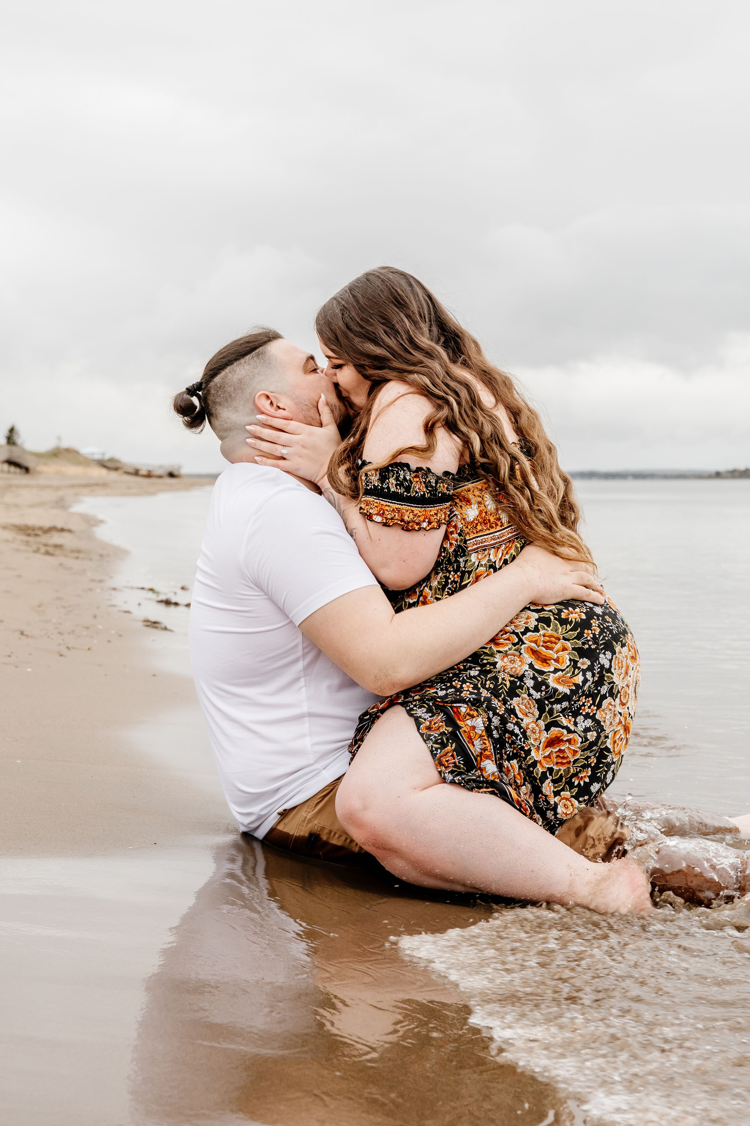 Couple kisses while sitting together in shallow beach waves during romantic engagement session.