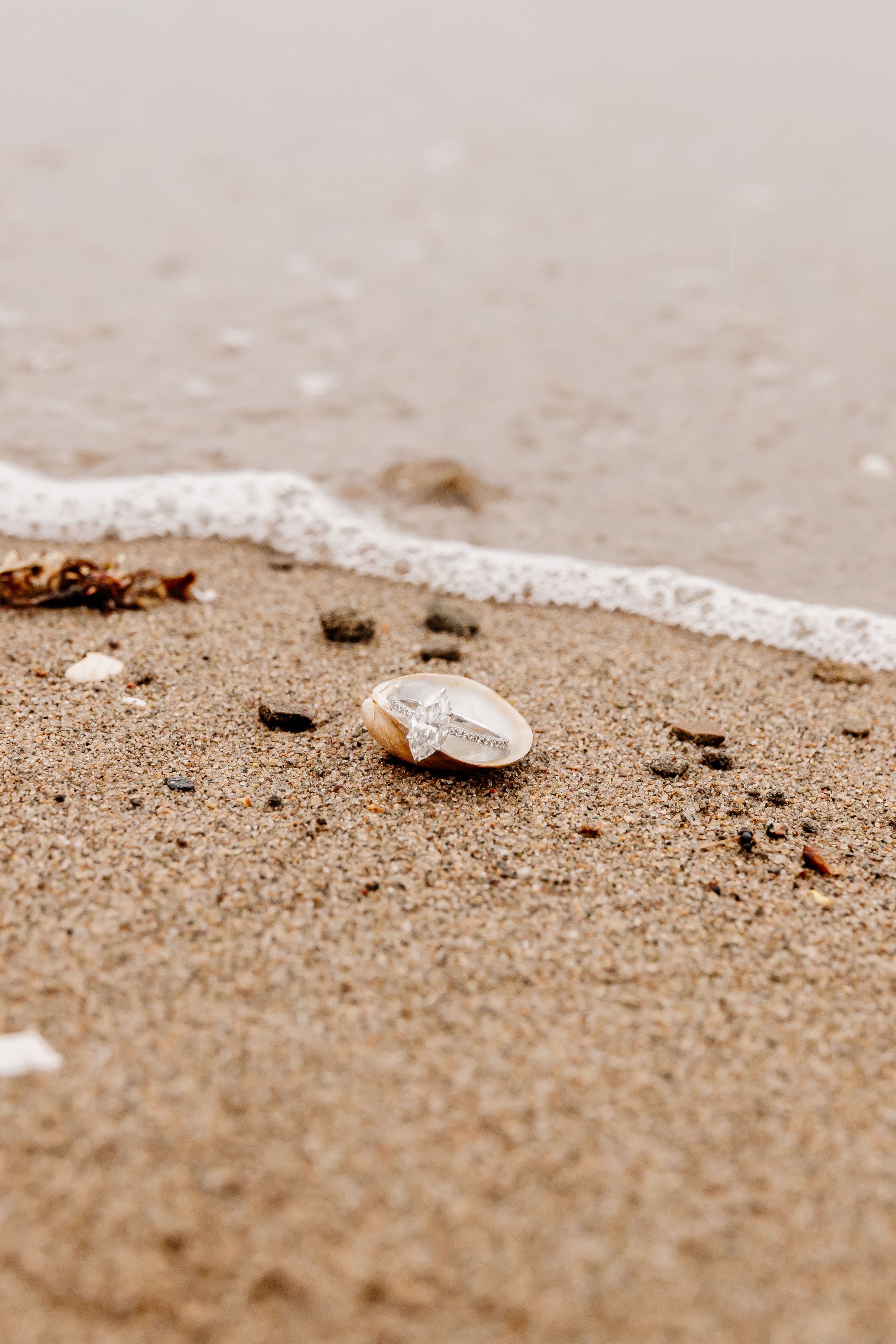 Diamond engagement ring resting in seashell on sandy beach with ocean waves nearby.