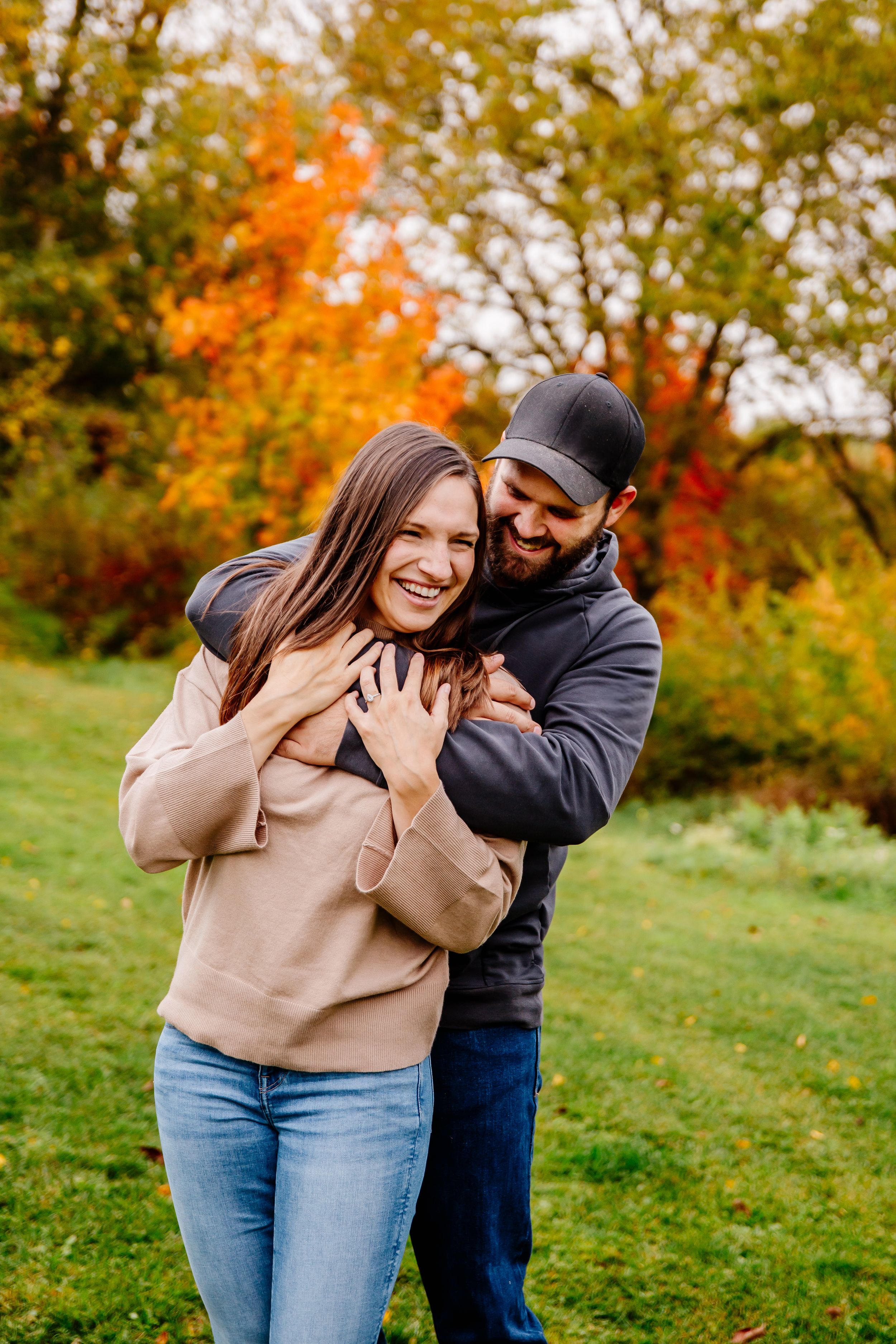 Happy couple embraces and laughs together outdoors with colorful autumn foliage in background.