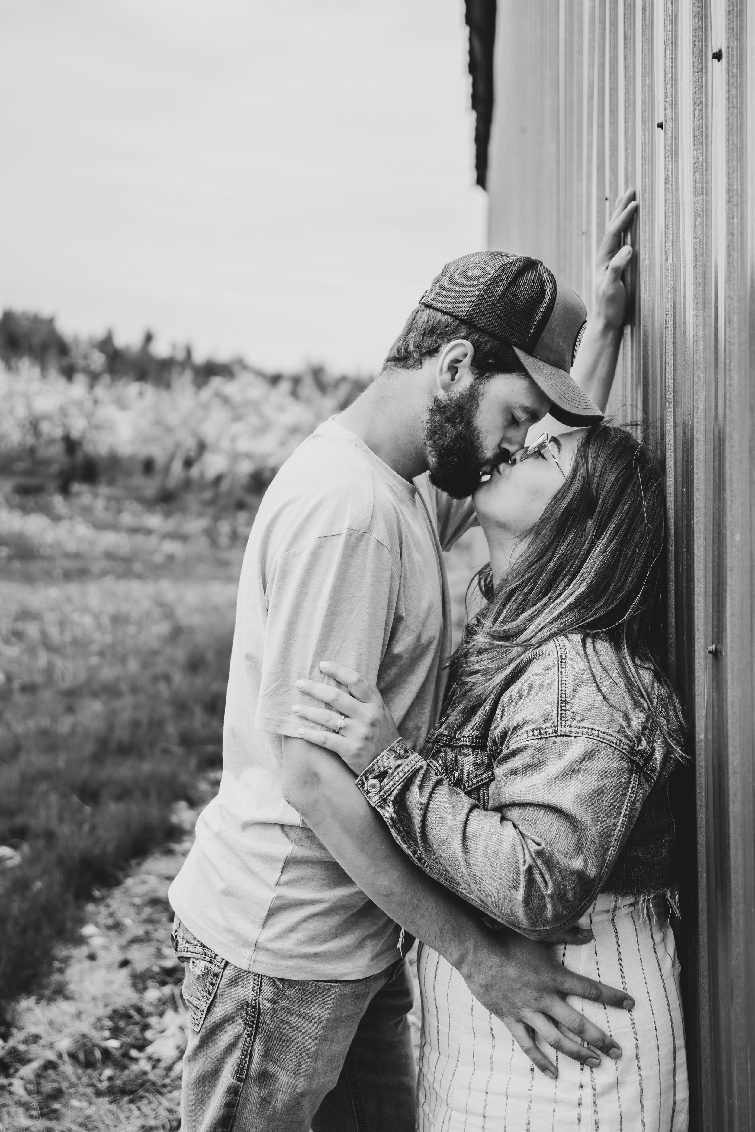 Black and white image of couple sharing a kiss against a barn wall in countryside.