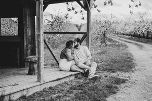 Black and white photo of couple sitting closely together on rustic porch near orchard.