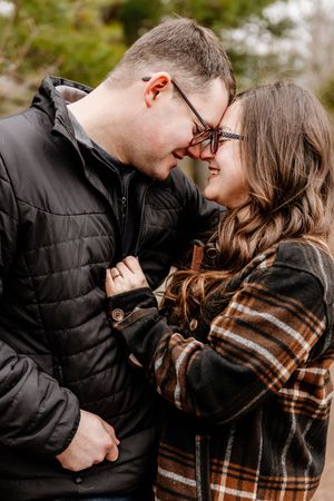 Smiling engaged couple in glasses touching foreheads outdoors in autumn forest setting.