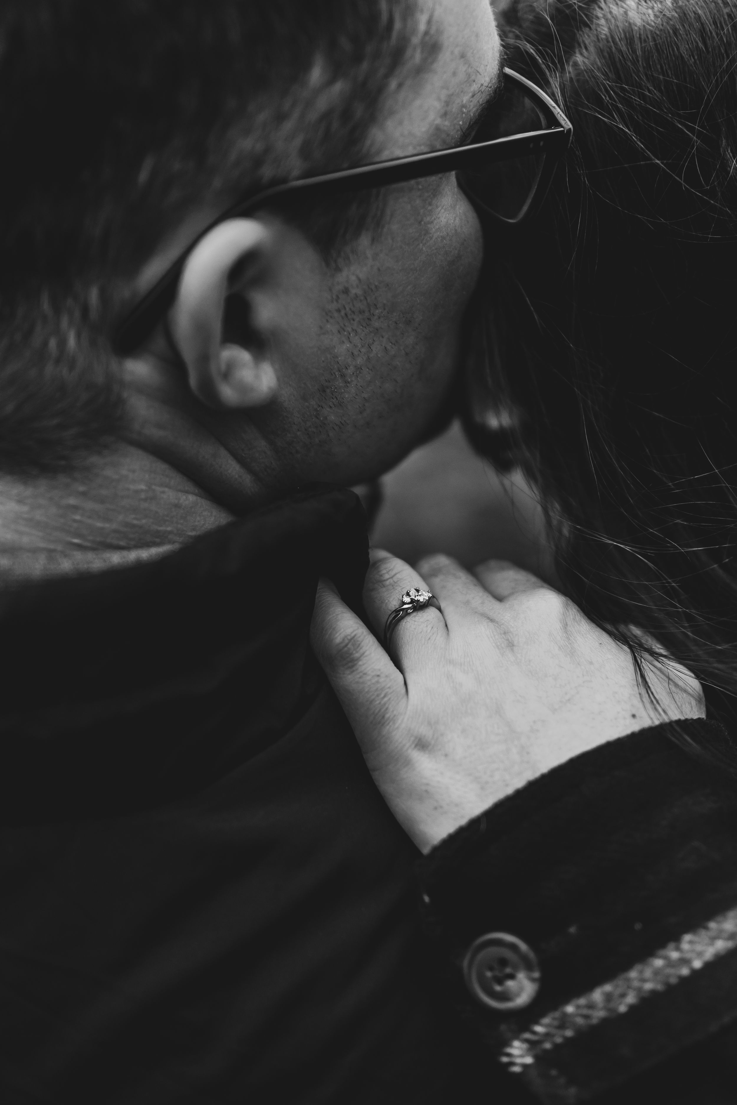 Black and white close-up of couple embracing, woman's hand with engagement ring resting on man's chest.
