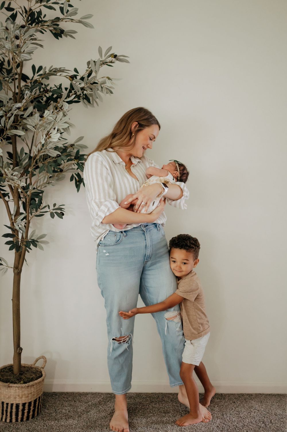 Mother in striped shirt holding newborn while toddler son hugs her legs near an olive tree.