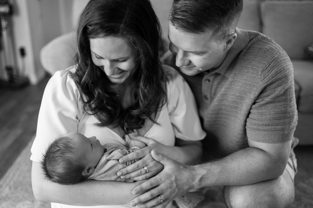 Black and white photo of smiling parents lovingly holding their newborn baby together.