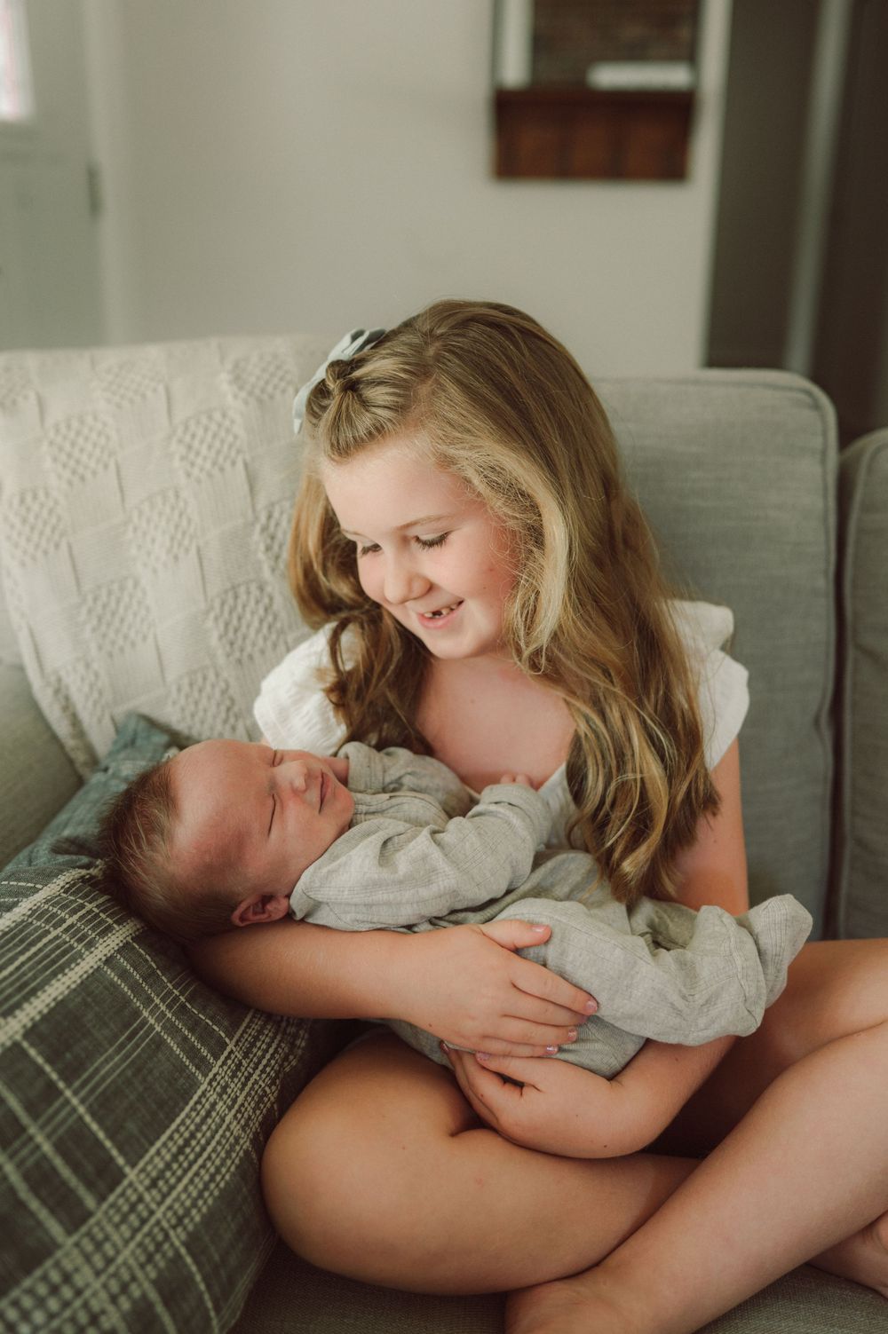 Smiling girl with long blonde hair holds sleeping newborn baby sibling on gray couch.