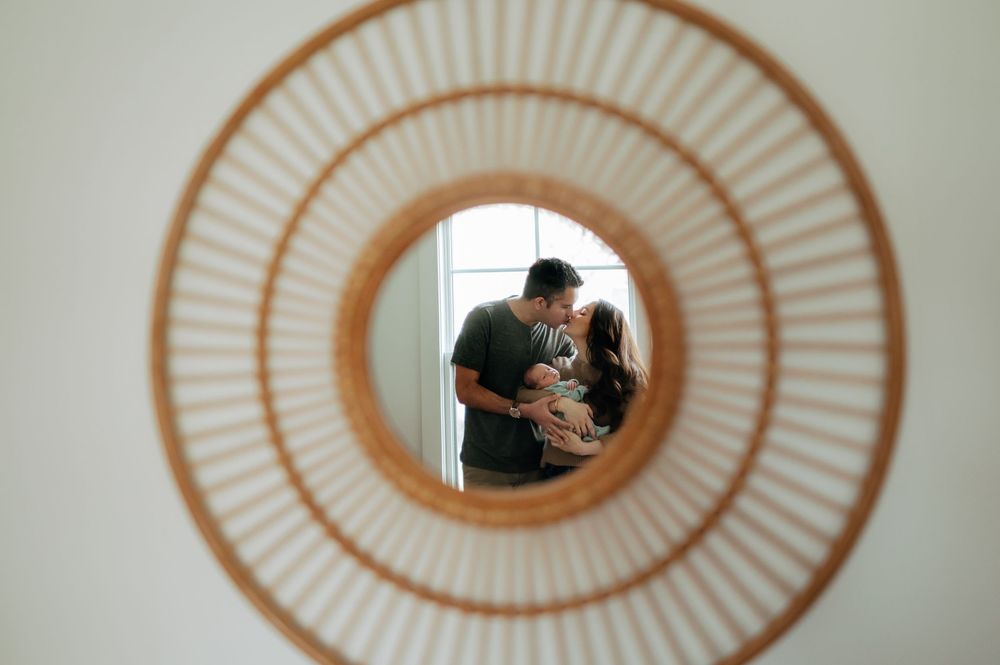Couple kissing while holding newborn baby reflected in a round rattan wall mirror.