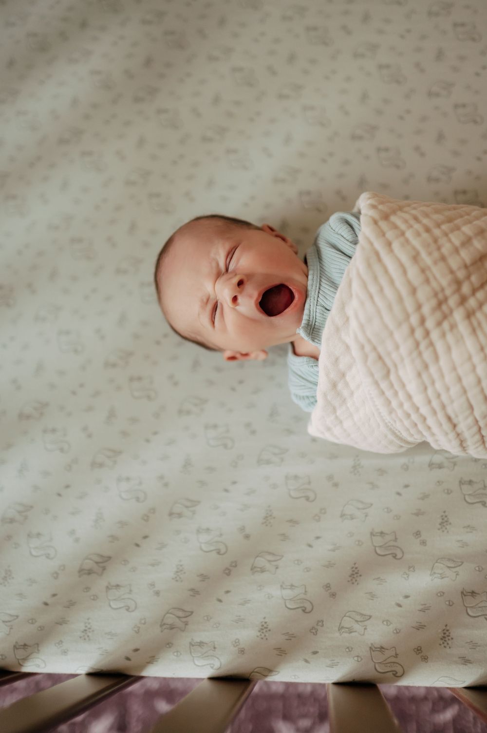 Swaddled newborn baby yawning while lying in a crib on a soft patterned sheet.