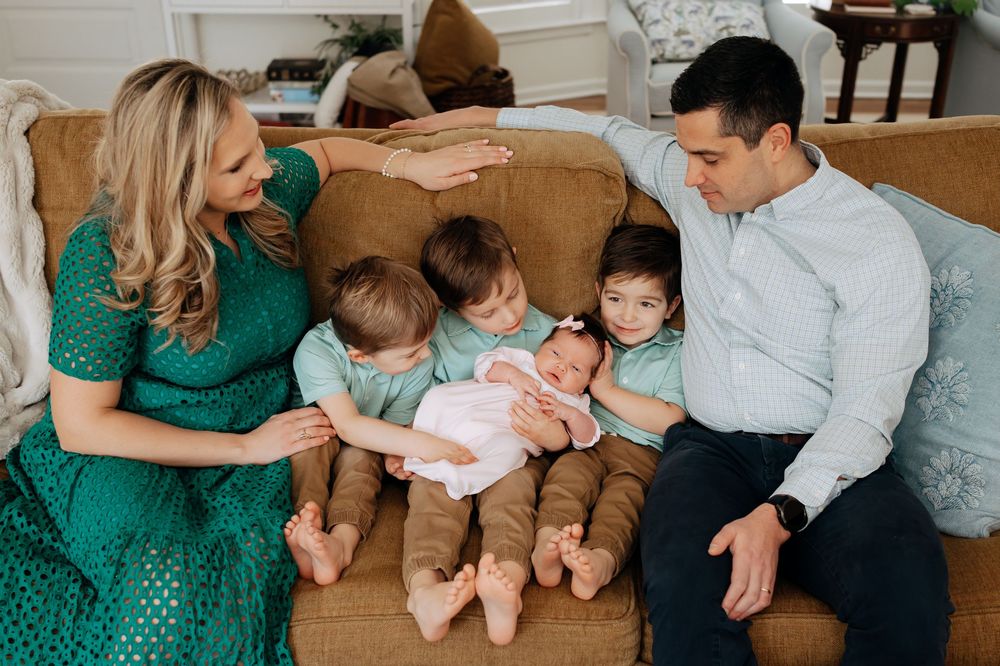 Family of five sitting on a couch, with three siblings lovingly gathered around their new baby.