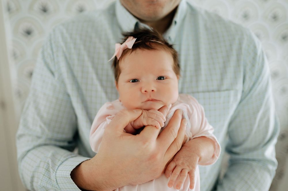 Father in a light blue shirt gently cradling his newborn baby girl wearing a pink bow headband.