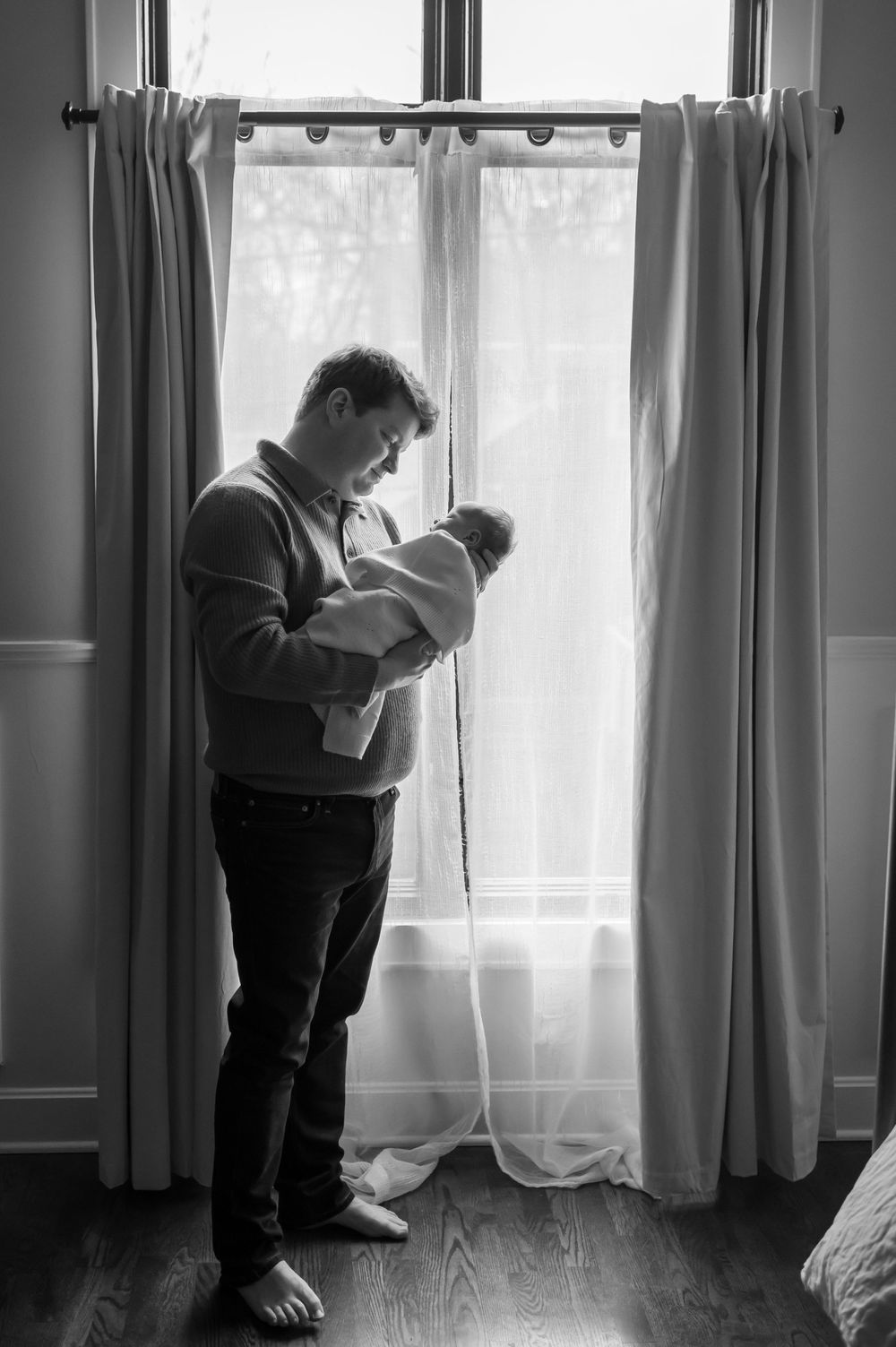 Black and white photo of a father standing by a window holding his newborn baby.