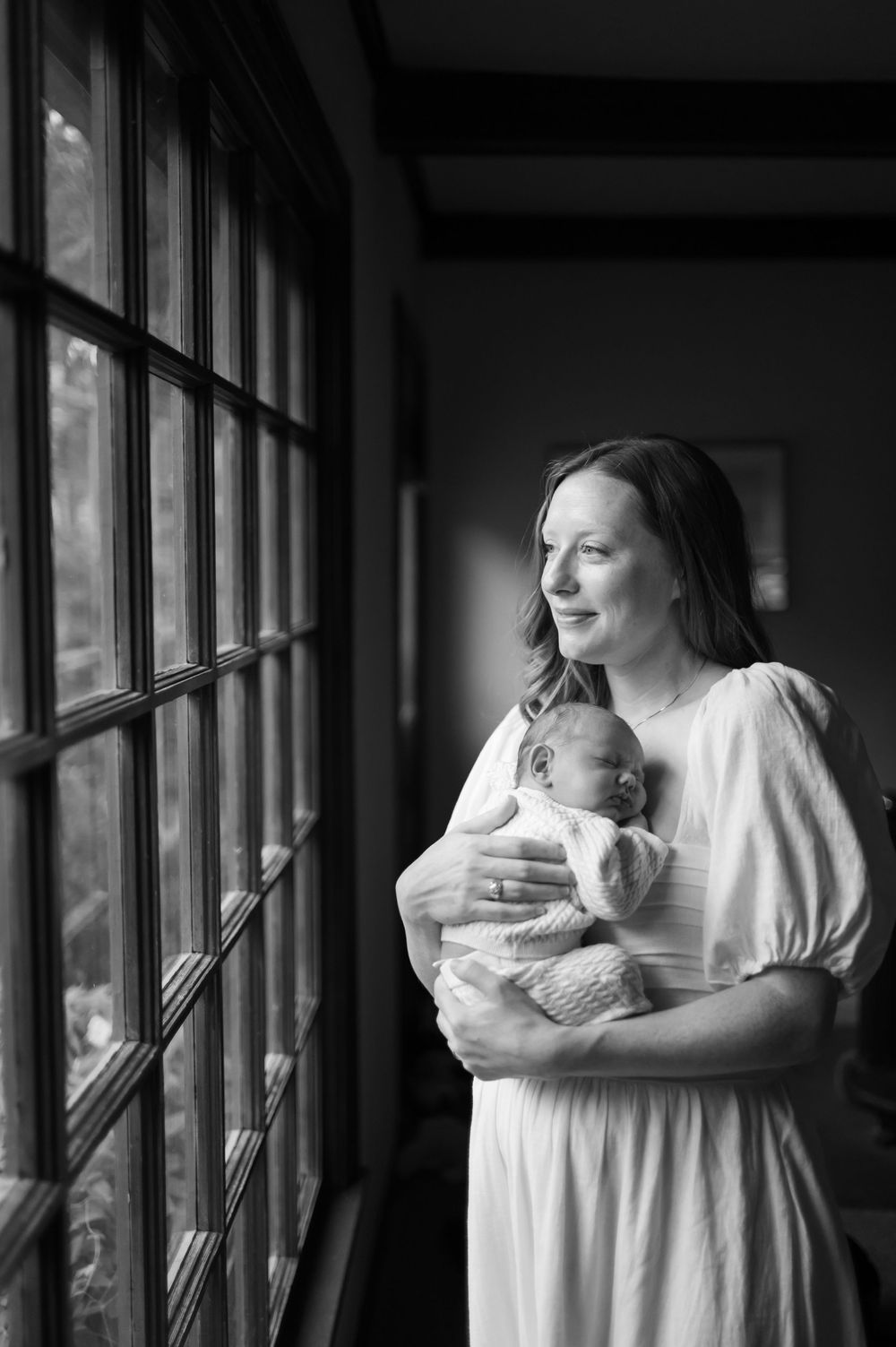 Black and white photo of a mother in a white dress holding her newborn while gazing out a window.