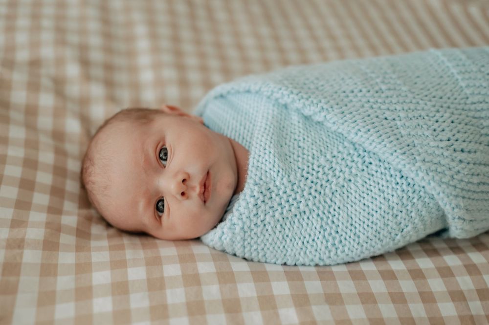 Newborn baby swaddled in a light blue knit blanket lying on a beige checkered blanket.