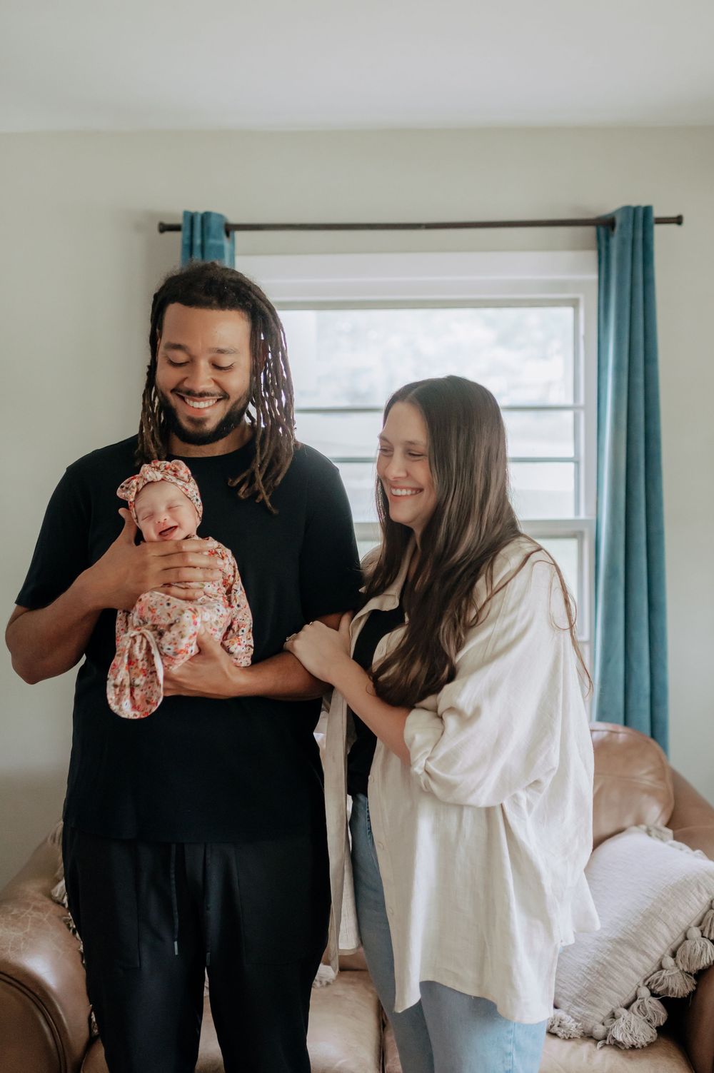 Smiling interracial couple standing indoors holding their newborn baby wrapped in a floral swaddle.