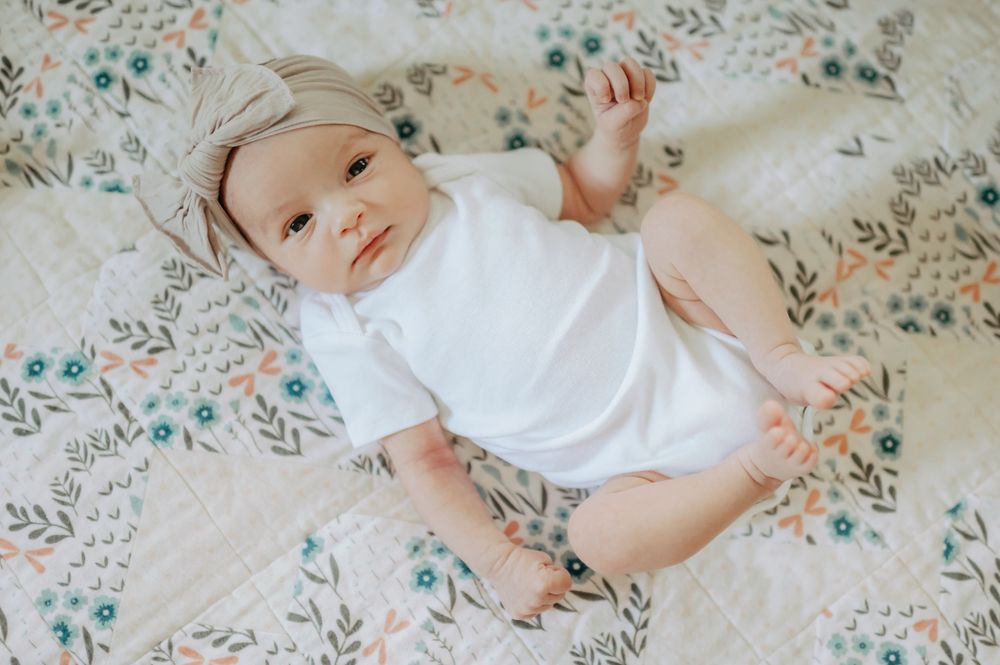 Adorable baby in white onesie and bow headband lying on floral quilt.