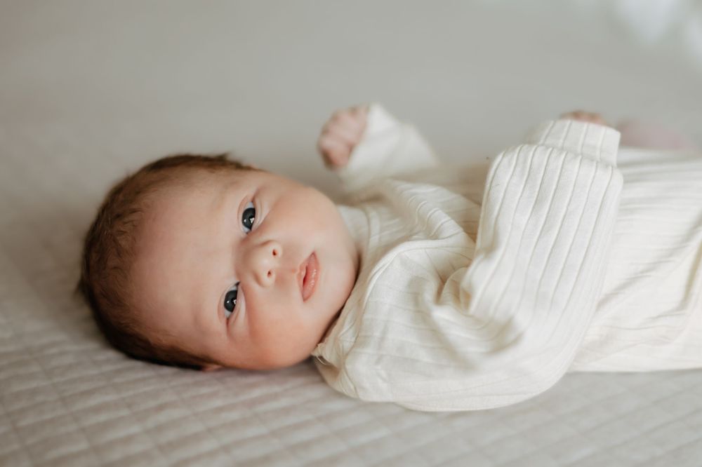 Newborn baby in a white knit outfit lying on a soft blanket with eyes open.