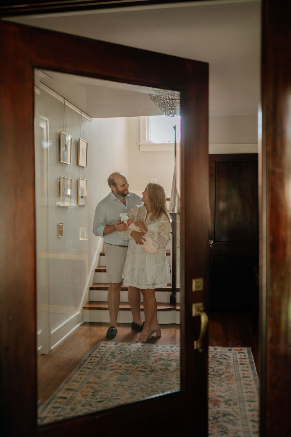 Couple with newborn baby photographed through doorway in warmly lit elegant home entryway.