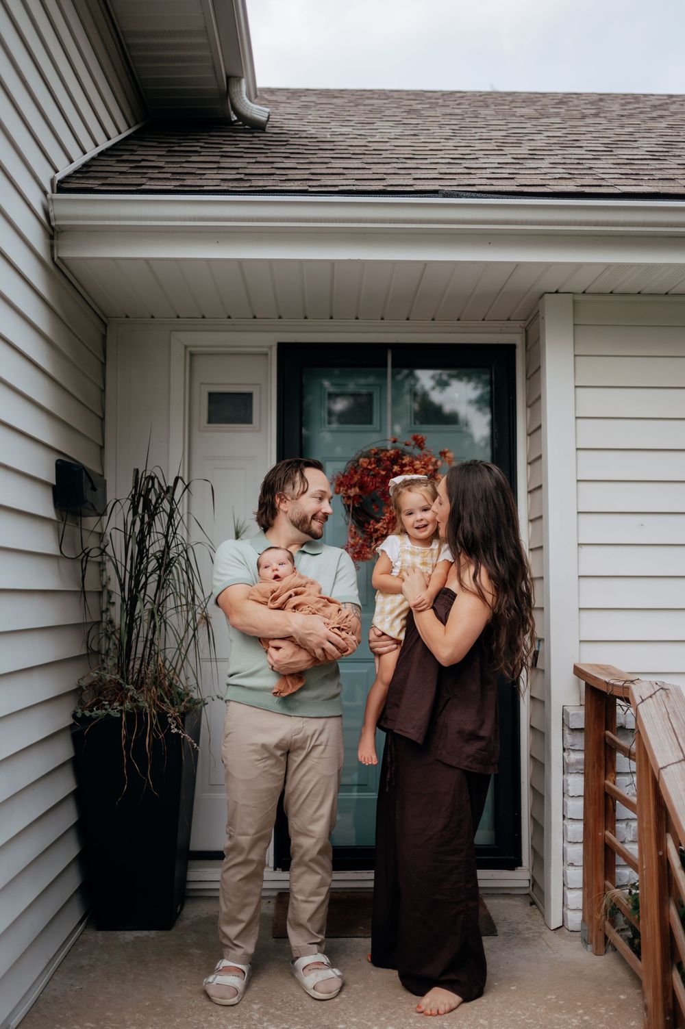Happy family of four standing at their front door, father holding newborn, mother holding toddler.