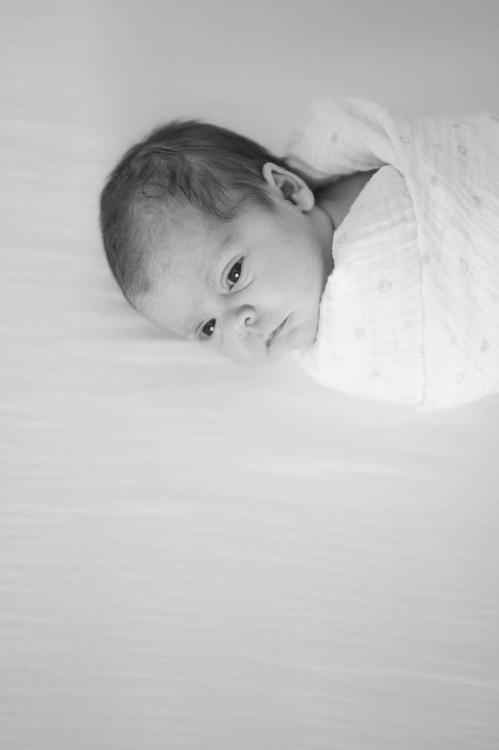 Black and white close-up portrait of alert newborn baby wrapped in white swaddle on soft surface.