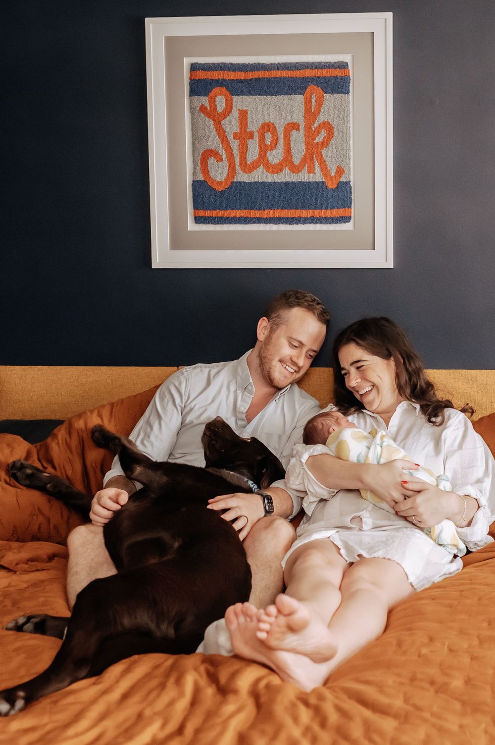 Happy new parents laughing on orange bed with newborn and black lab beneath a family name sign.
