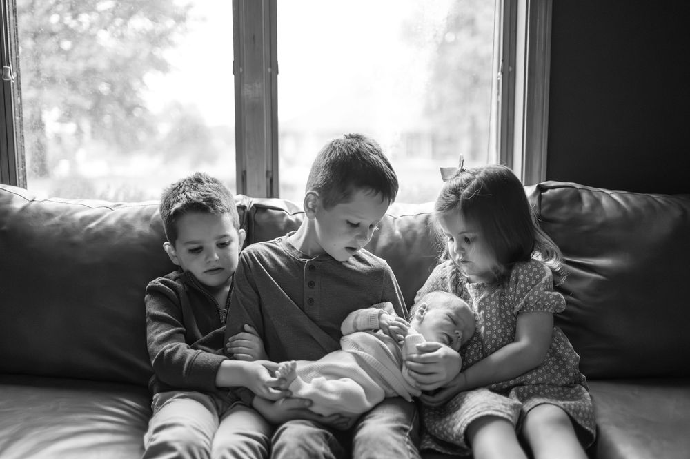 Black and white photo of three siblings sitting on a couch together holding a newborn baby.