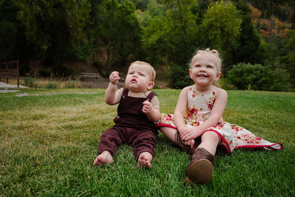 Two young siblings sitting together on grass with colorful autumn trees behind.