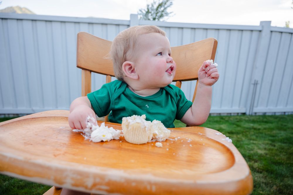 Happy toddler in green onesie sitting in wooden high chair eating smash cake outdoors.