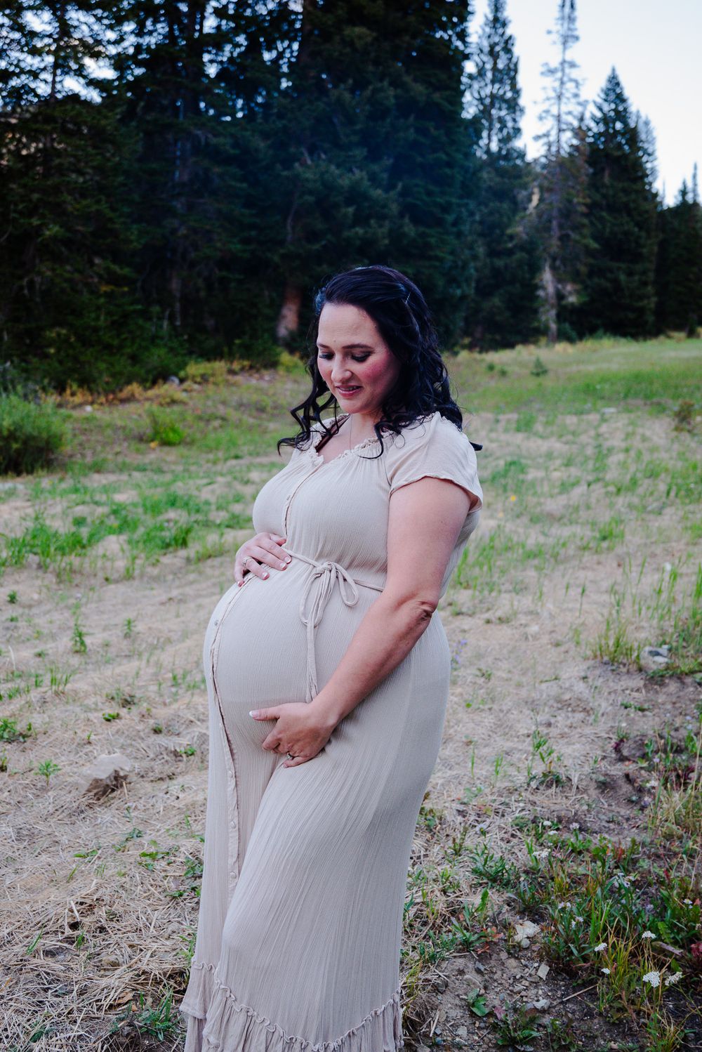 Pregnant woman in beige maxi dress cradling belly in forest meadow setting.
