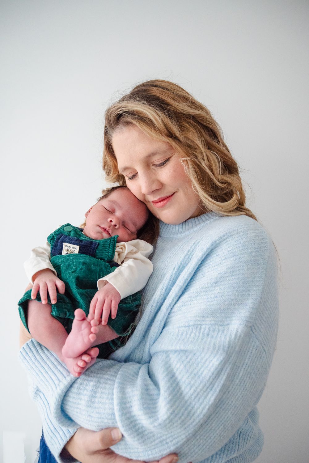 A smiling mother in a blue sweater cradles her sleeping newborn baby against her cheek.