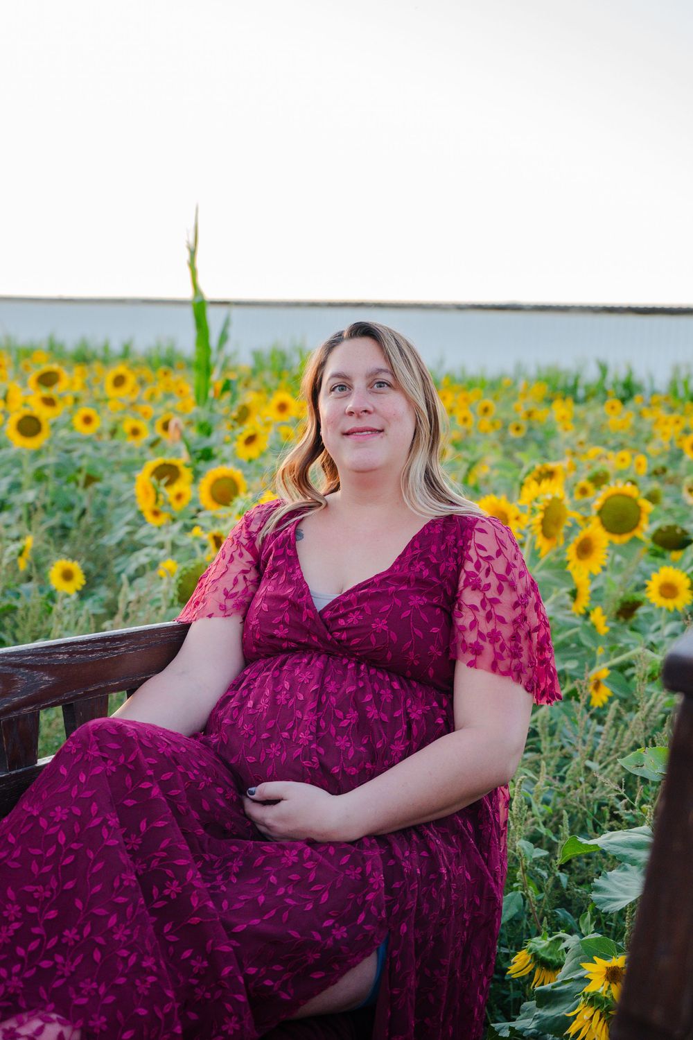 Smiling pregnant woman in burgundy dress sitting on bench surrounded by sunflowers.
