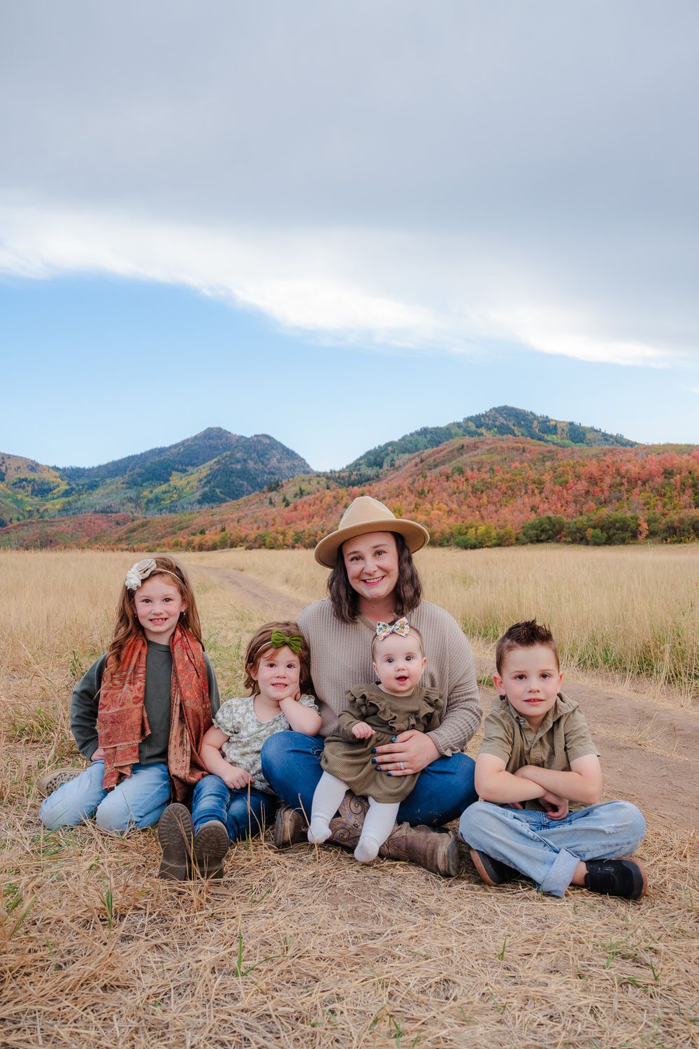 A mother in a tan hat sits with her four young children in a golden field with colorful autumn mountains behind.