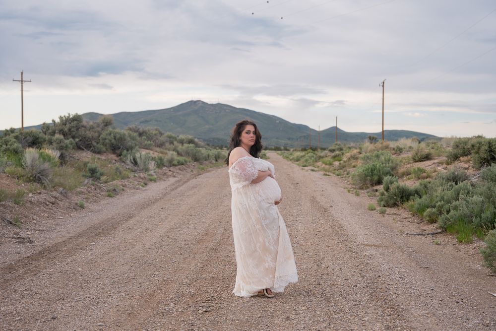 Pregnant woman in white lace maternity gown standing on rural dirt road with mountains in background.
