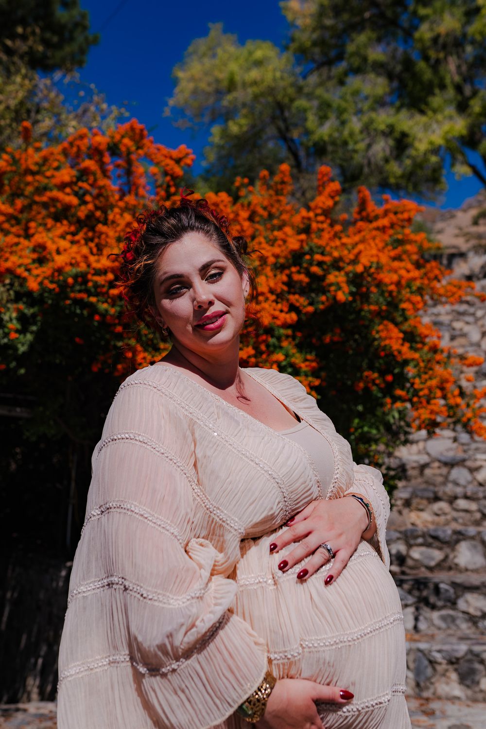 Smiling pregnant woman in cream dress holding belly before bright orange flowers outdoors.