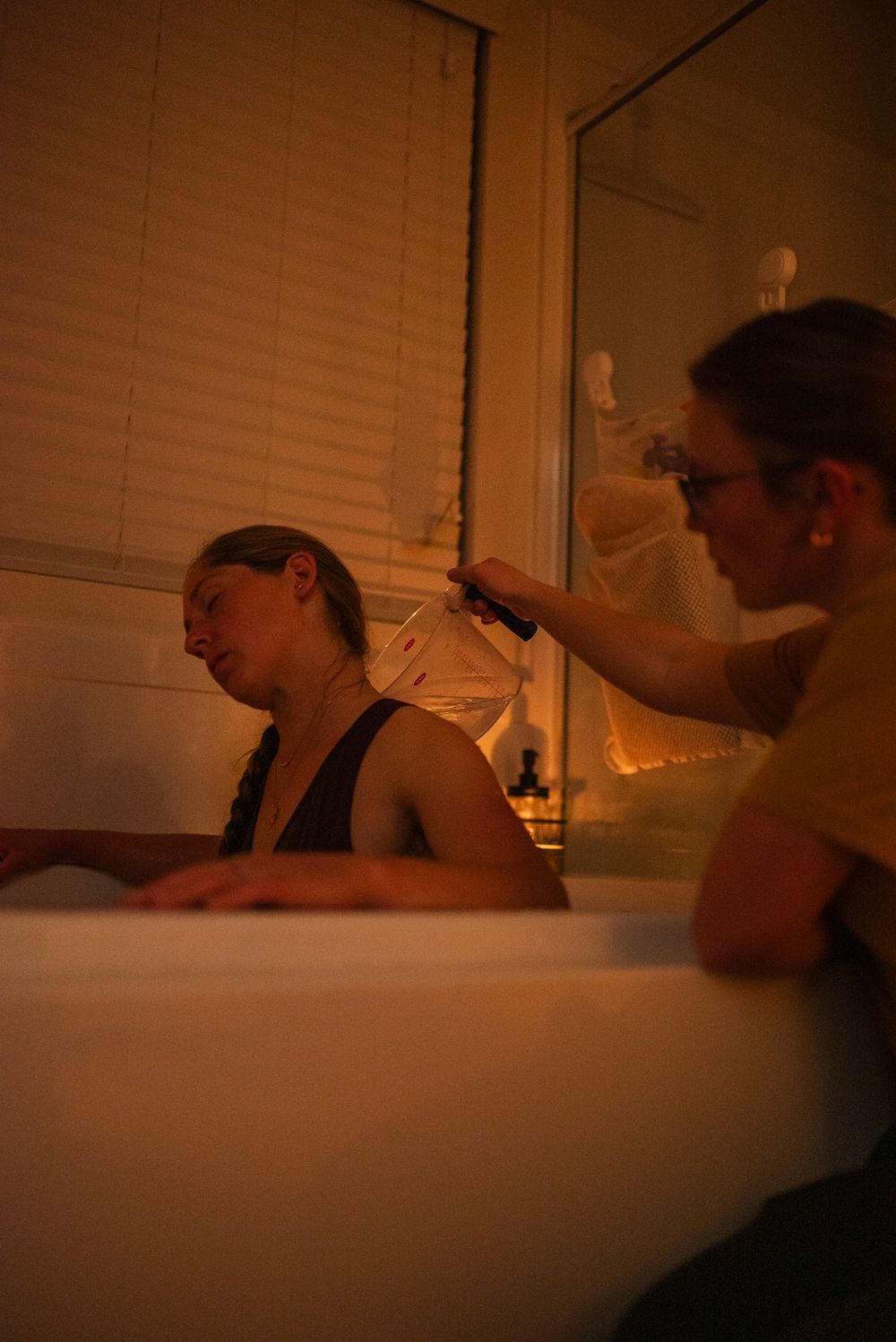 A woman relaxes in a birth tub while a doula pours warm water over her back during labor.