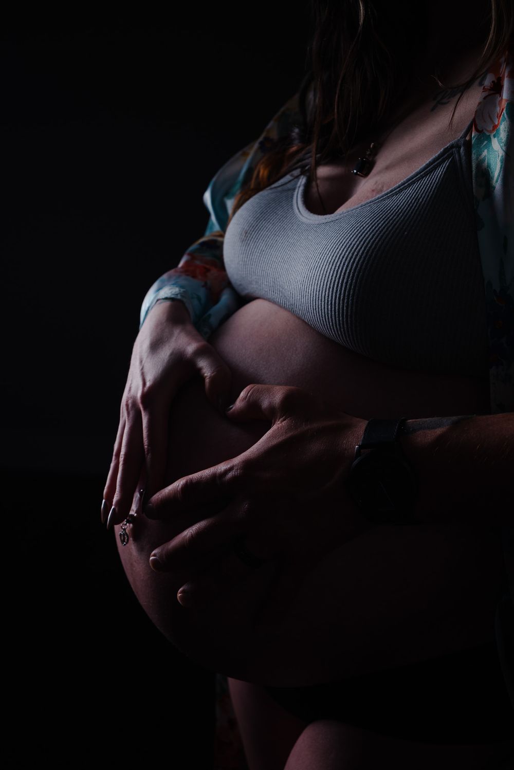 Dramatic low-key maternity photo of couple's hands tenderly holding pregnant belly in grey crop top.