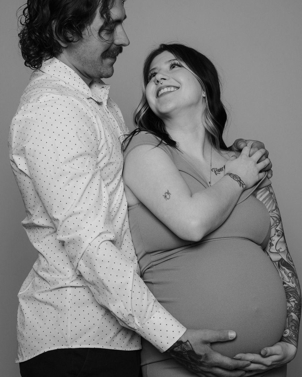 Black and white maternity photo of laughing couple embracing, woman with tattoos holding pregnant belly.