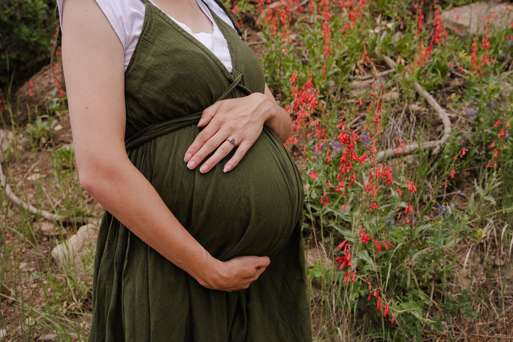 Pregnant woman in olive green dress holding baby bump near vibrant red wildflowers in nature.