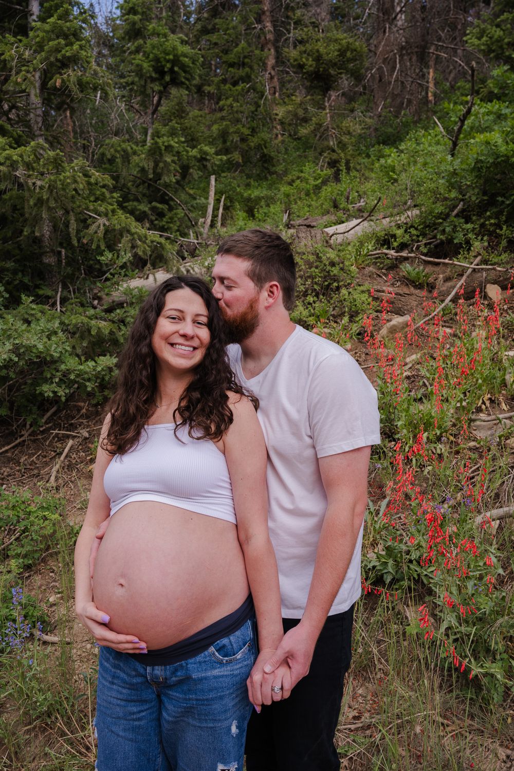 Man kissing pregnant woman on cheek as she smiles and holds her baby bump in a forest setting.