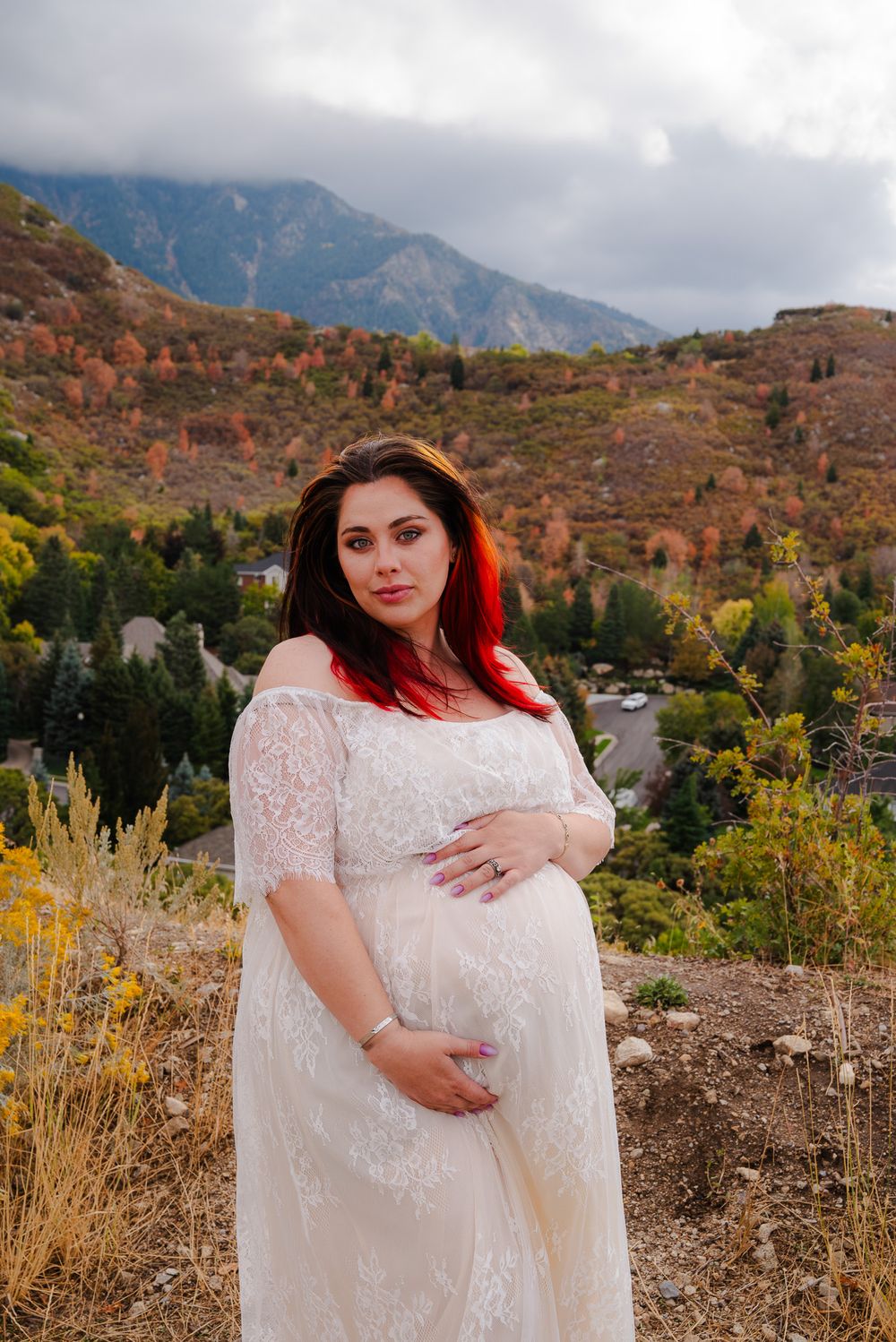 Pregnant woman in white lace dress cradles baby bump against colorful autumn mountain landscape.