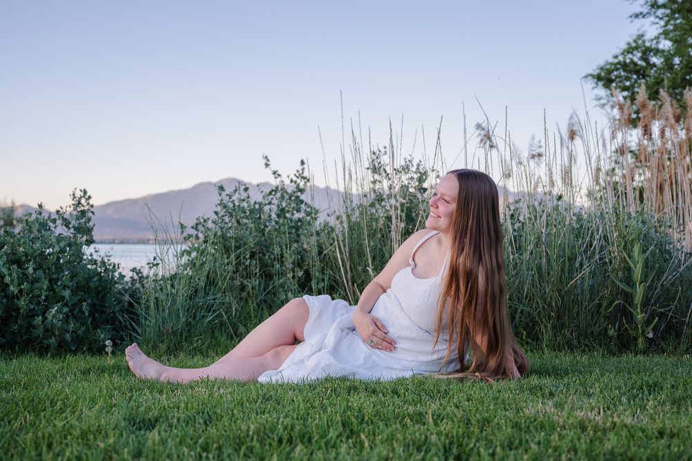 Smiling pregnant woman in white dress sitting barefoot on grass near lake at dusk.