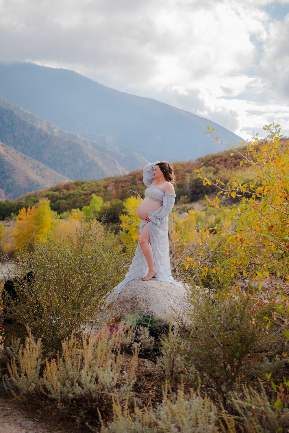 Pregnant woman in sheer white gown stands on boulder gazing at stunning autumn mountain landscape.