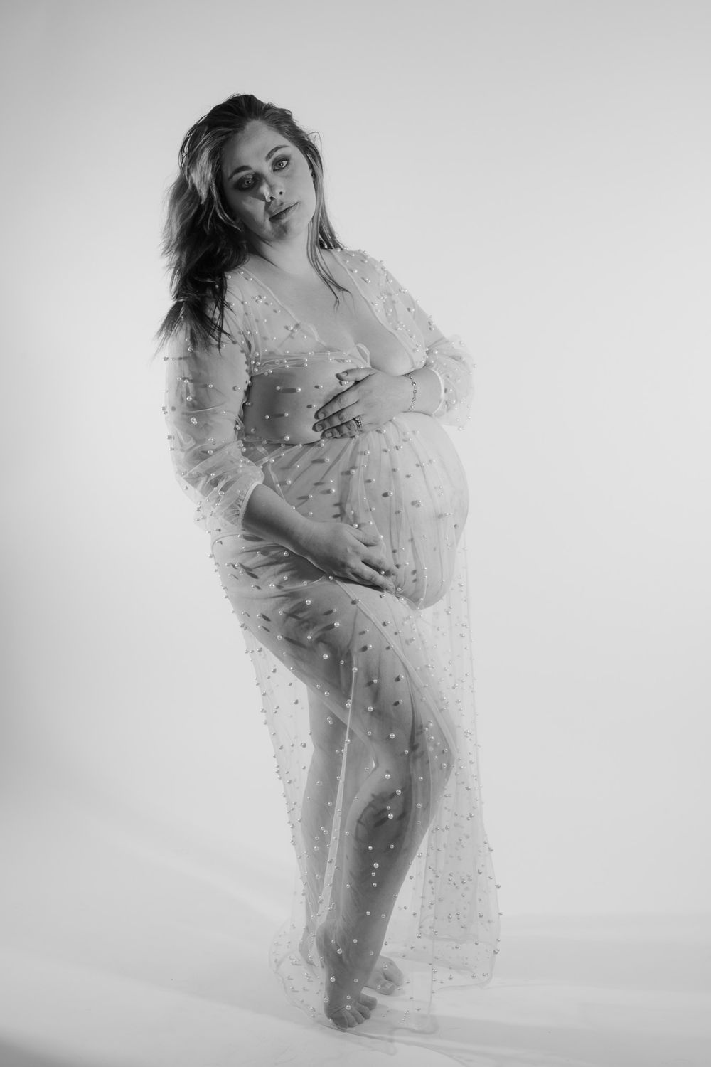 Black and white photo of pregnant woman in sheer pearl-embellished gown posing in studio.