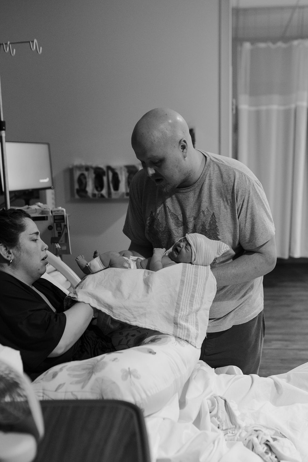 Black and white photo of new parents in hospital room admiring their swaddled newborn baby.