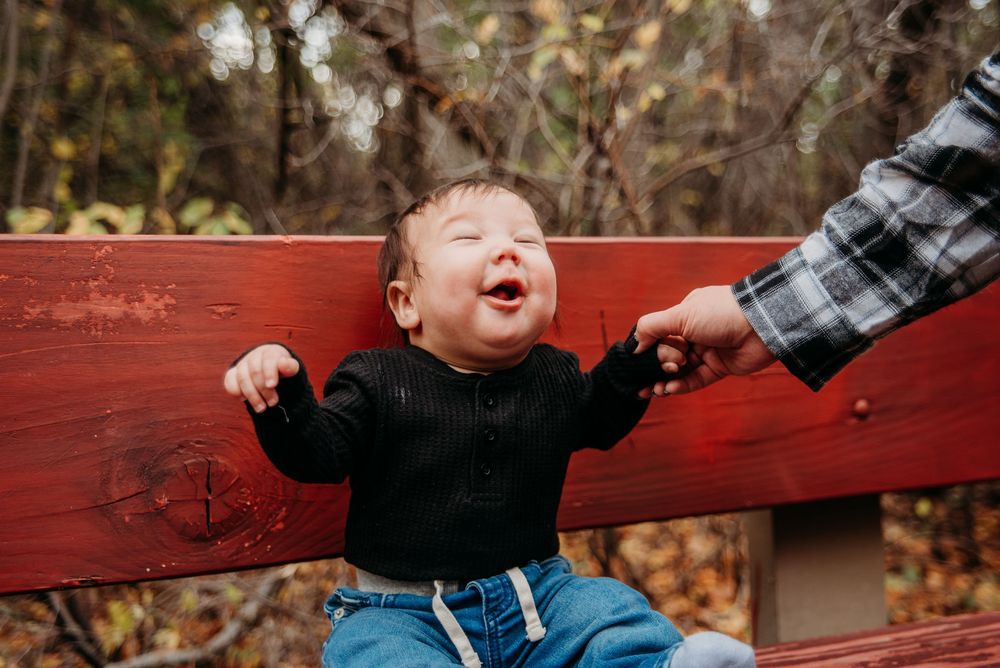 A joyful baby in a black shirt sits on a red bench holding a parent's hand outdoors.