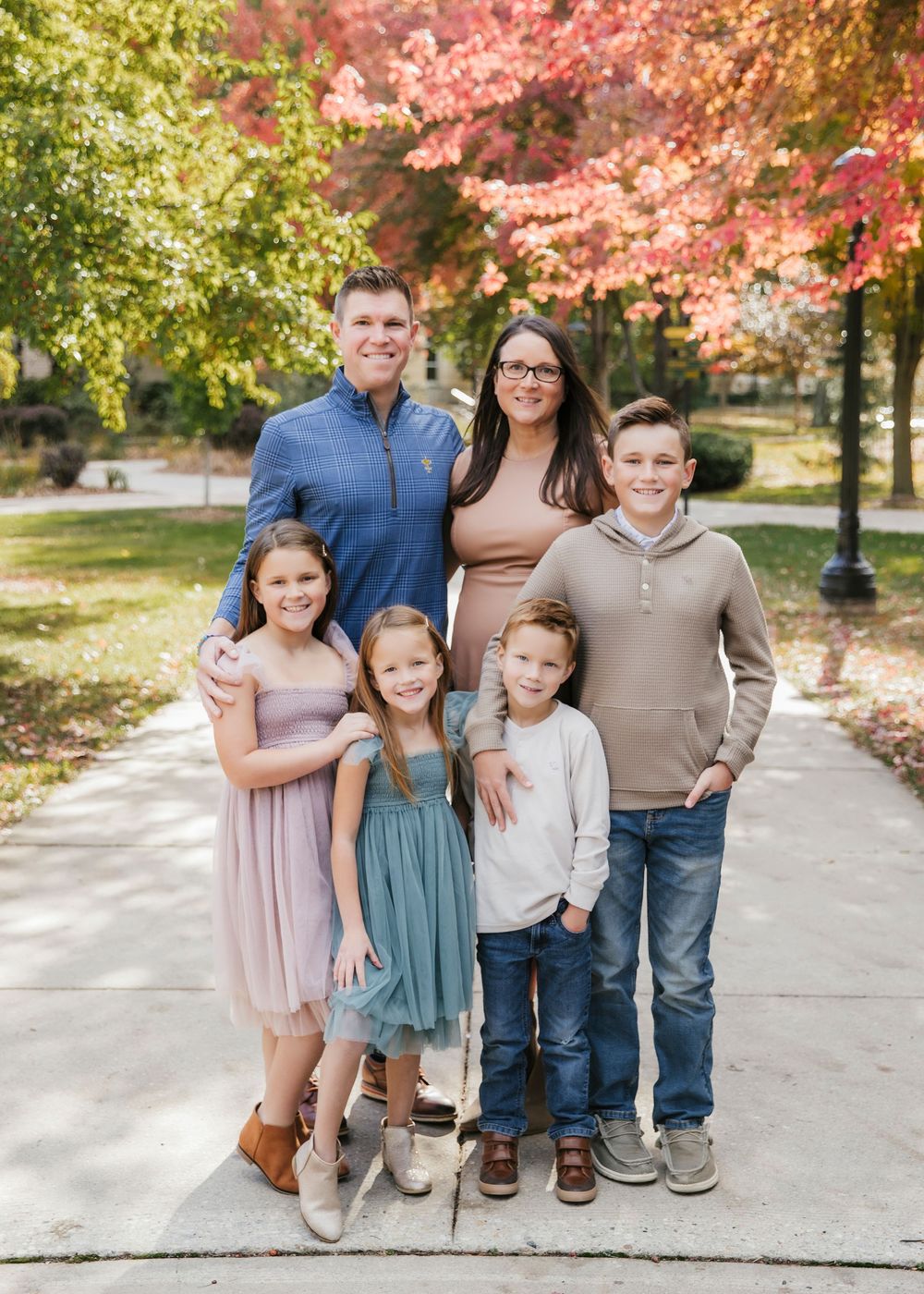 Family of six poses together on sidewalk with autumn foliage in background.