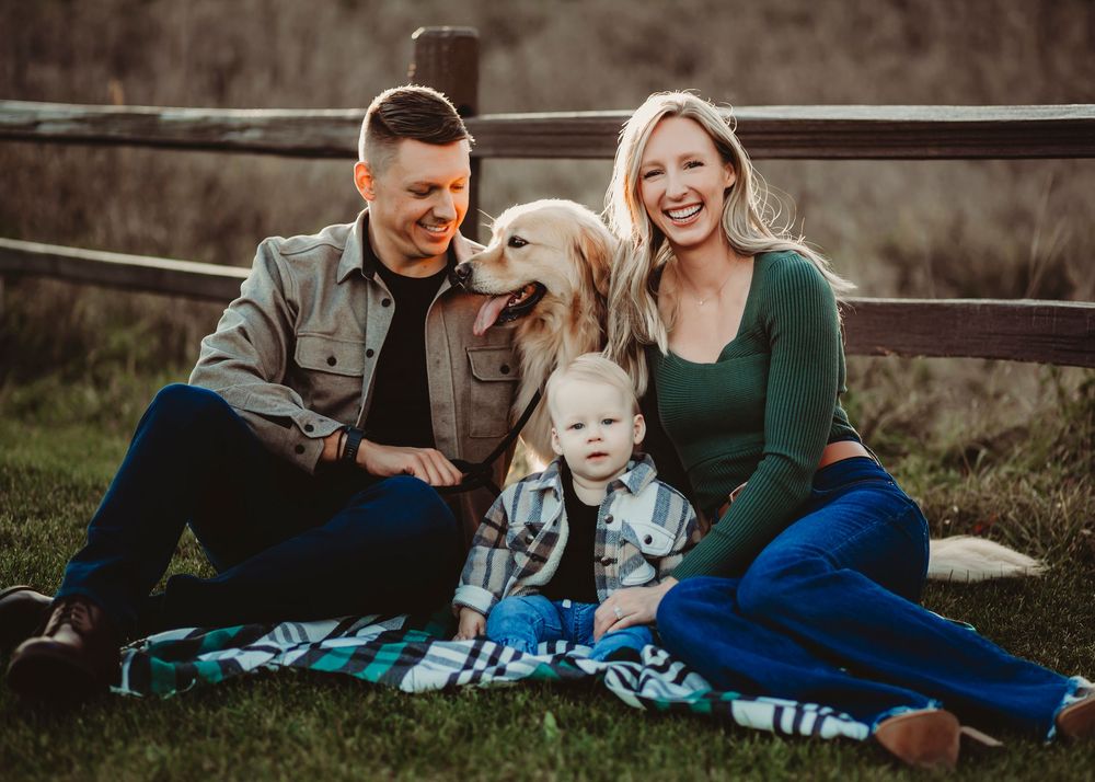 Family with golden retriever dog sits on blanket by rustic fence at sunset.